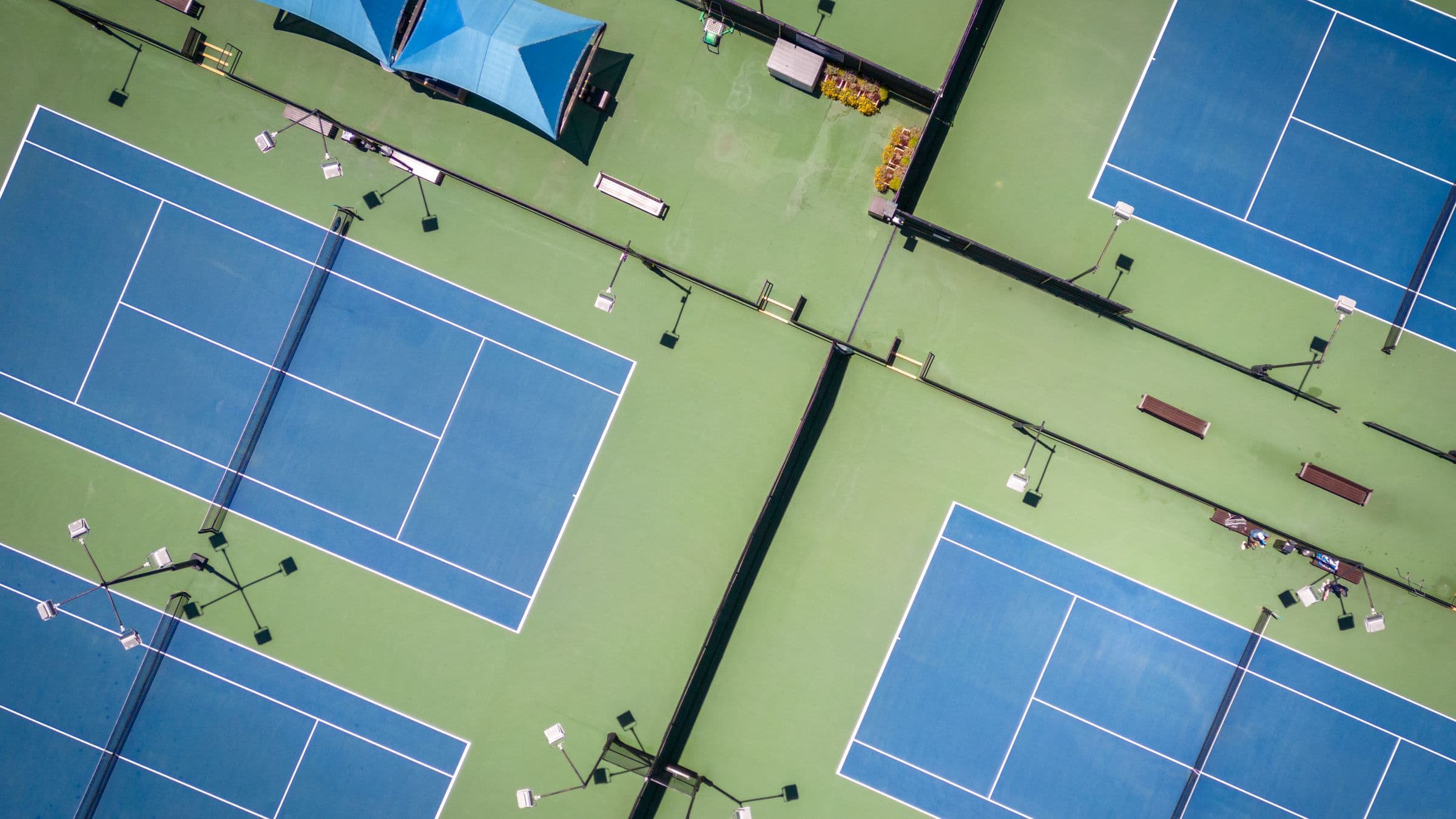 Four hard courts photographed from directly overhead, blue playing surfaces marked out on a green surround, with floodlight masts, benches and a shade canopy between them. Nobody is playing.