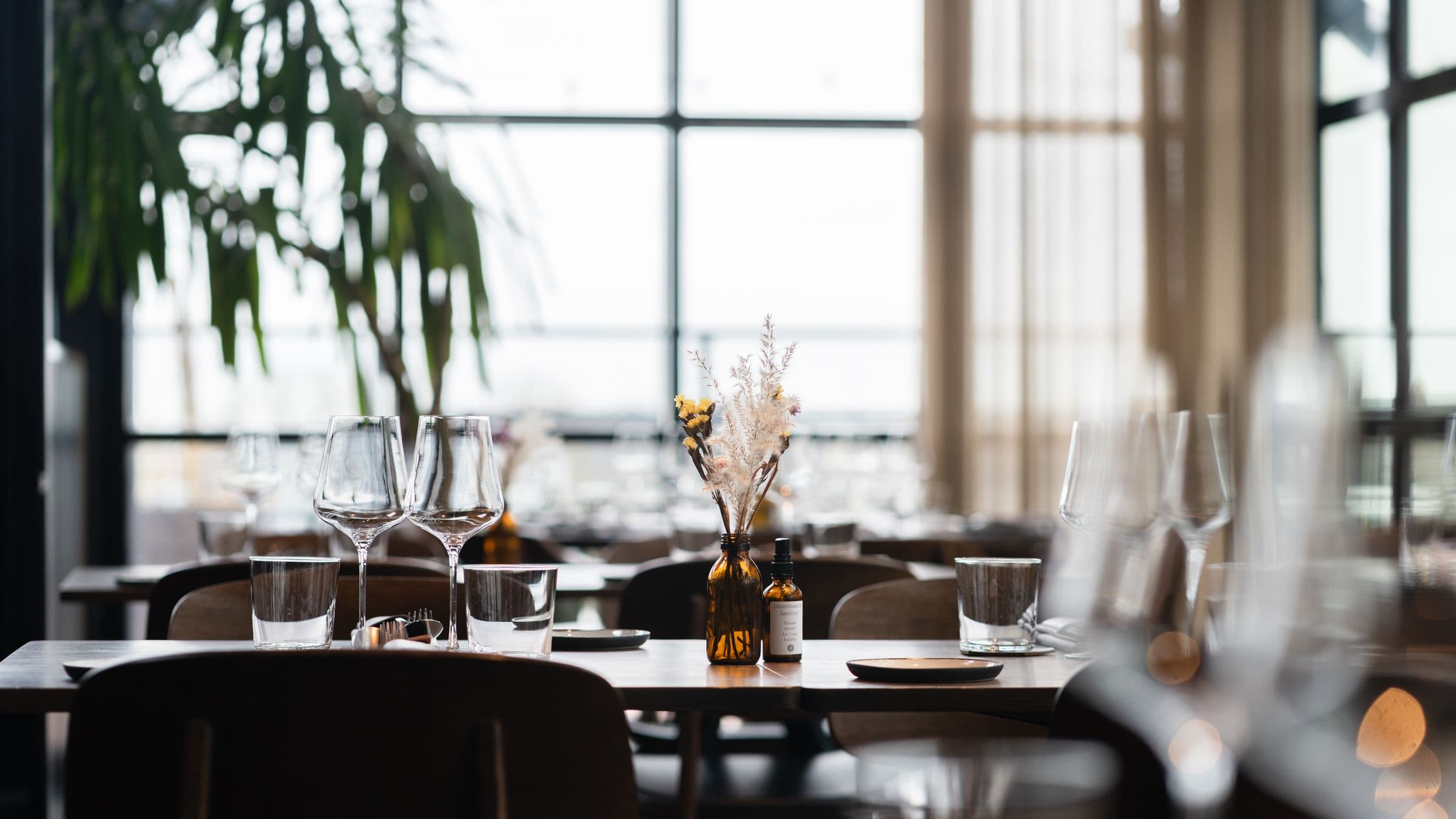 An empty dining room in morning light: one table laid with wine glasses, tumblers and a small vase, with tall windows and more set tables behind it.