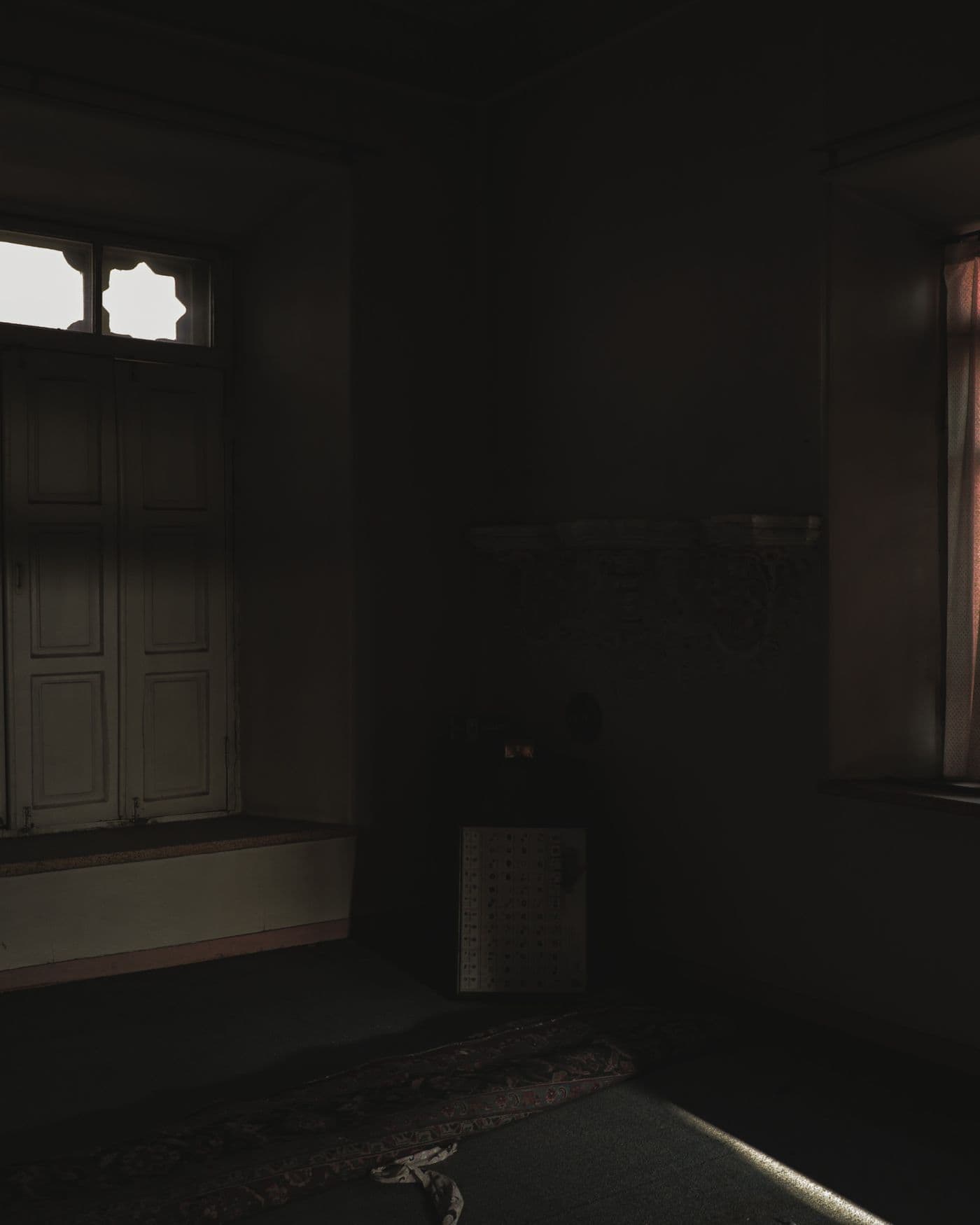 An empty room in near-darkness, its only light falling through one shuttered window.